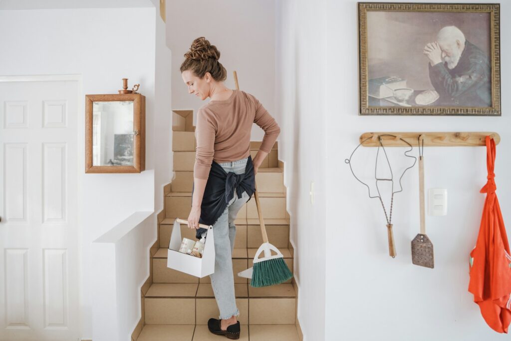 A woman carrying a cleaning caddy and broom up tiled stairs in a home