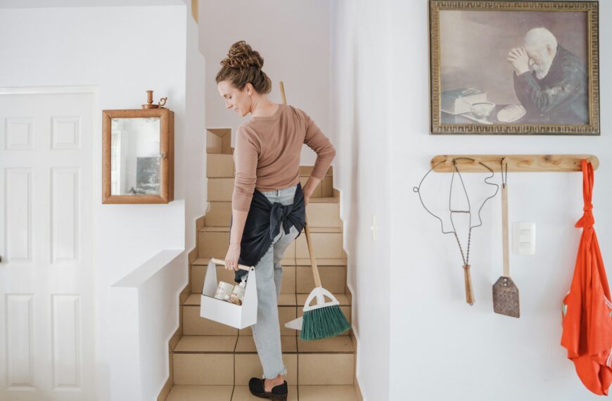 A woman carrying a cleaning caddy and broom up tiled stairs in a home