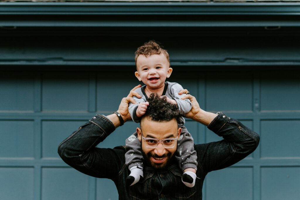 An adult with glasses carrying a baby on his shoulders against a blue door