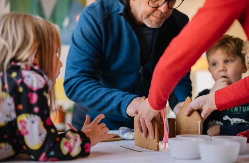 two person making gingerbread house and three children watching them