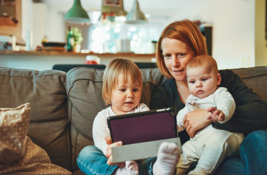 two babies and woman sitting on sofa while holding baby and watching on tablet
