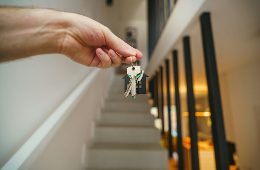 A hand holding keys with a house-shaped keychain in front of a staircase