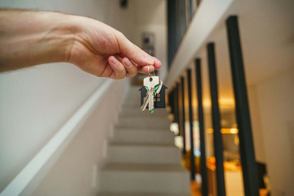 A hand holding keys with a house-shaped keychain in front of a staircase
