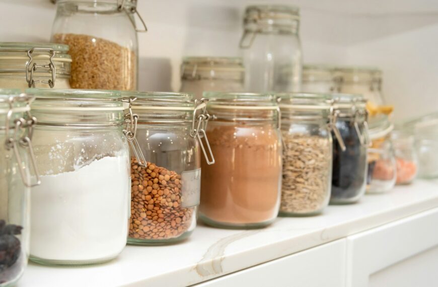 Rows of glass storage jars filled with flour, lentils, grains and other dry goods on a white pantry shelf.
