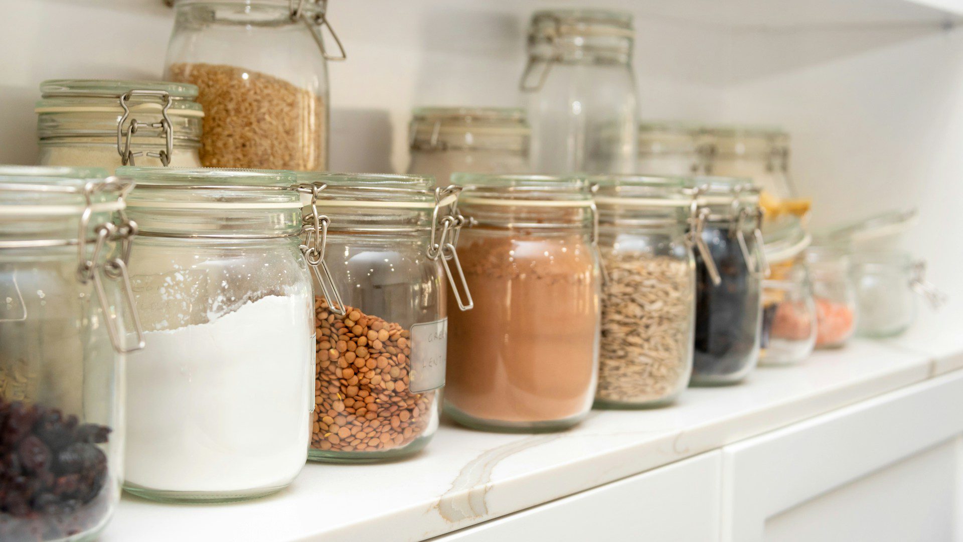 Rows of glass storage jars filled with flour, lentils, grains and other dry goods on a white pantry shelf.