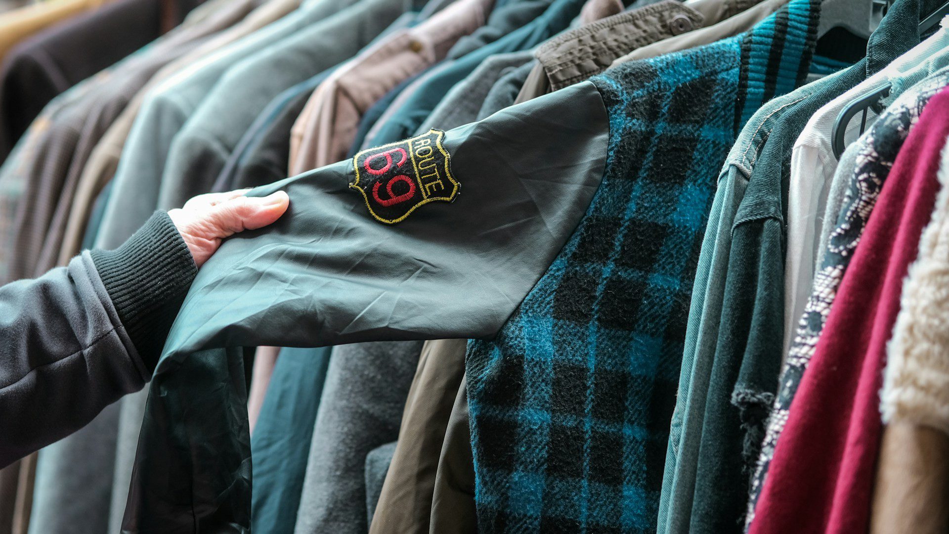 A hand holds a shirt on a crowded rack of secondhand clothes.