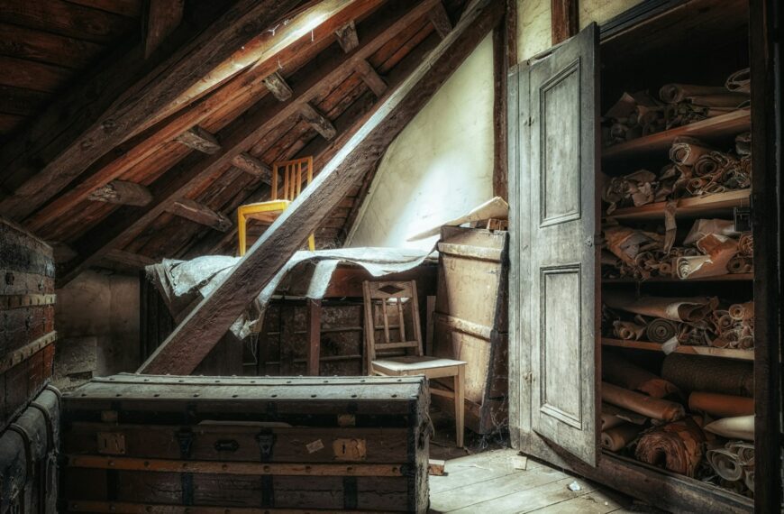 An attic under a sloped wooden roof with an old trunk, furniture and a cupboard of stored rolls (Unsplash photo by Peter Herrmann).