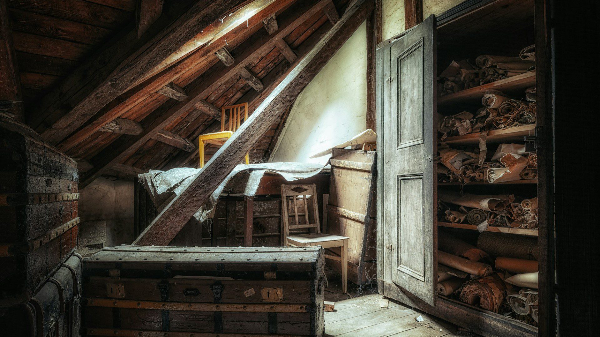 An attic under a sloped wooden roof with an old trunk, furniture and a cupboard of stored rolls (Unsplash photo by Peter Herrmann).