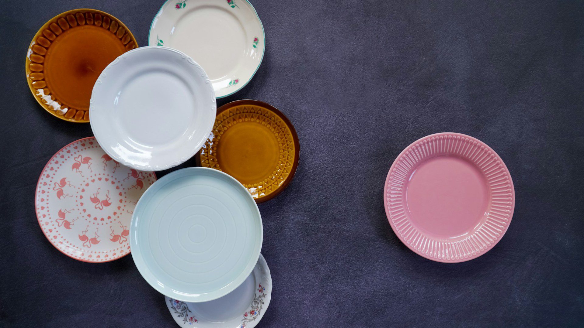 Several vintage plates in amber, pastel blue, pink and floral patterns on a dark surface (Unsplash photo by Karolina Kołodziejczak).