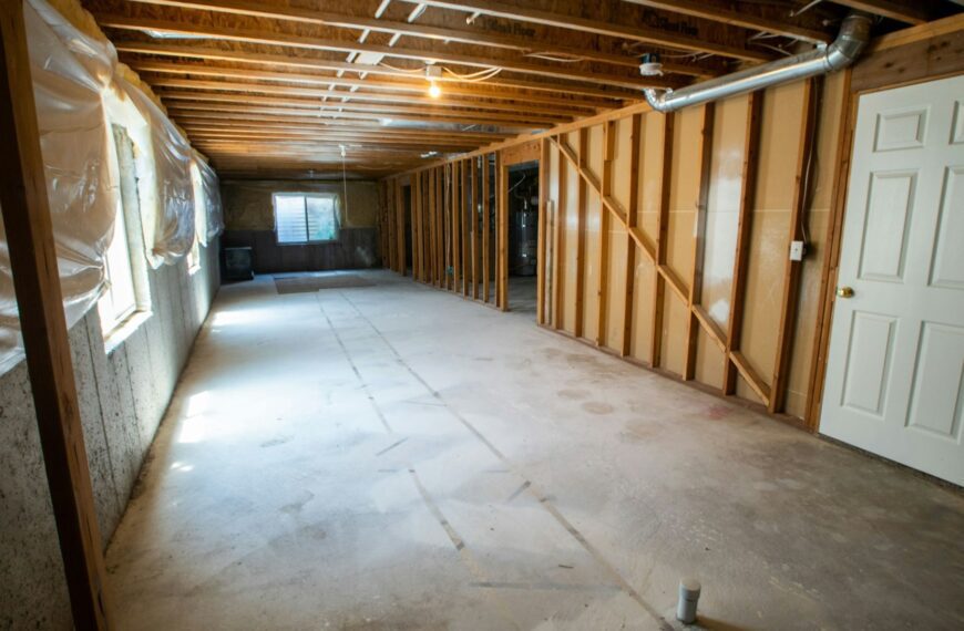 An unfinished basement with exposed beams, a concrete floor and a white door (Unsplash photo by Real Twin Photos).