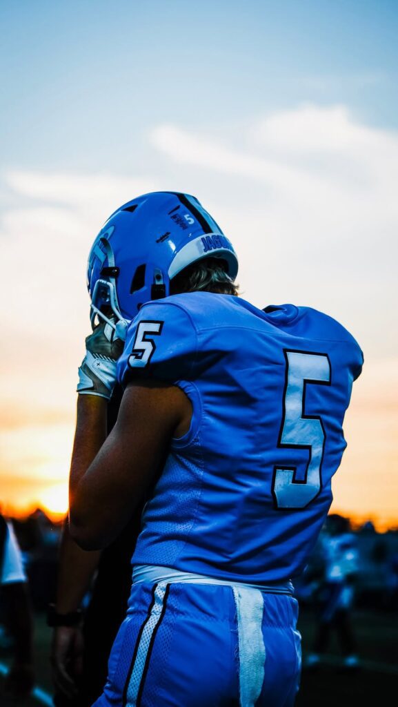 A football player in a blue number 5 jersey and helmet, seen from behind at sunset.